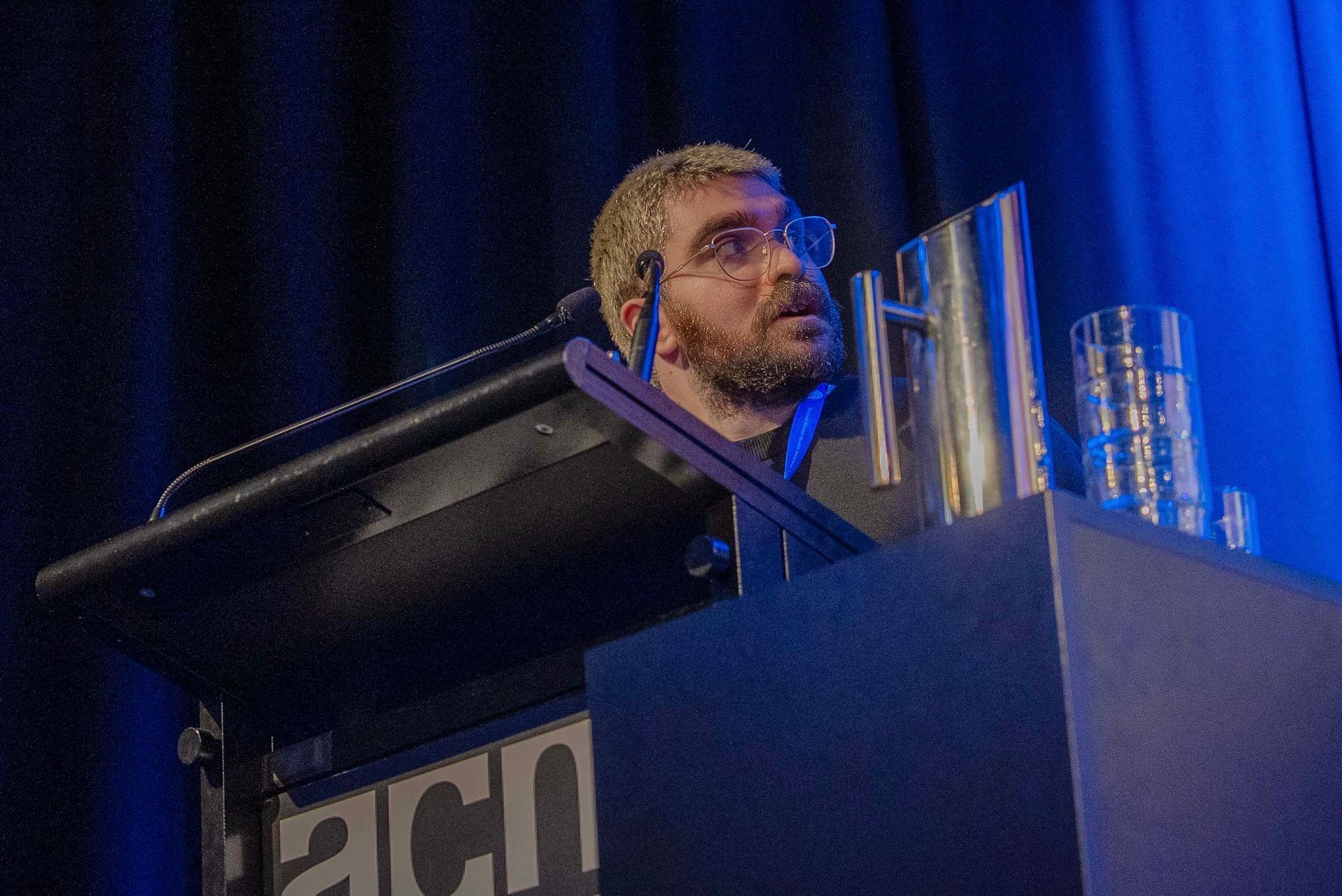 Man speaking at a podium with microphones under blue stage lighting
