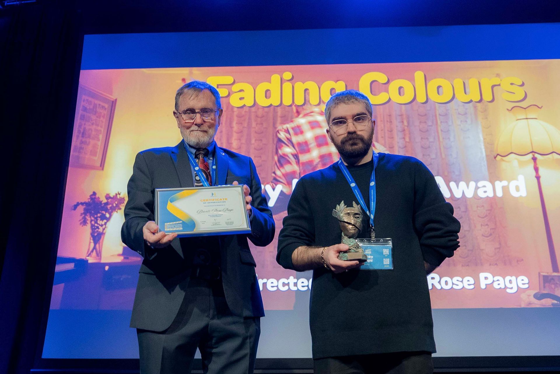 Two men on stage holding an award and certificate in front of a colorful event backdrop