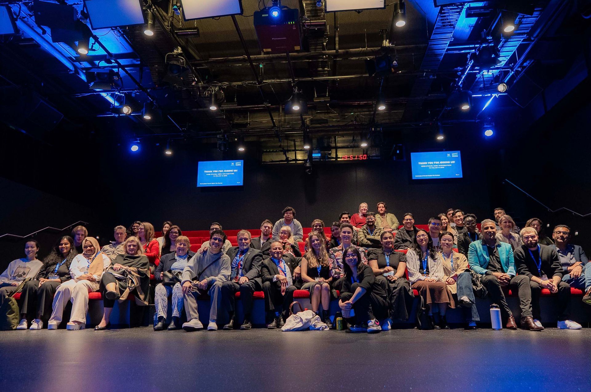 Large group seated on a stage under blue lights in a theater, with two small screens overhead