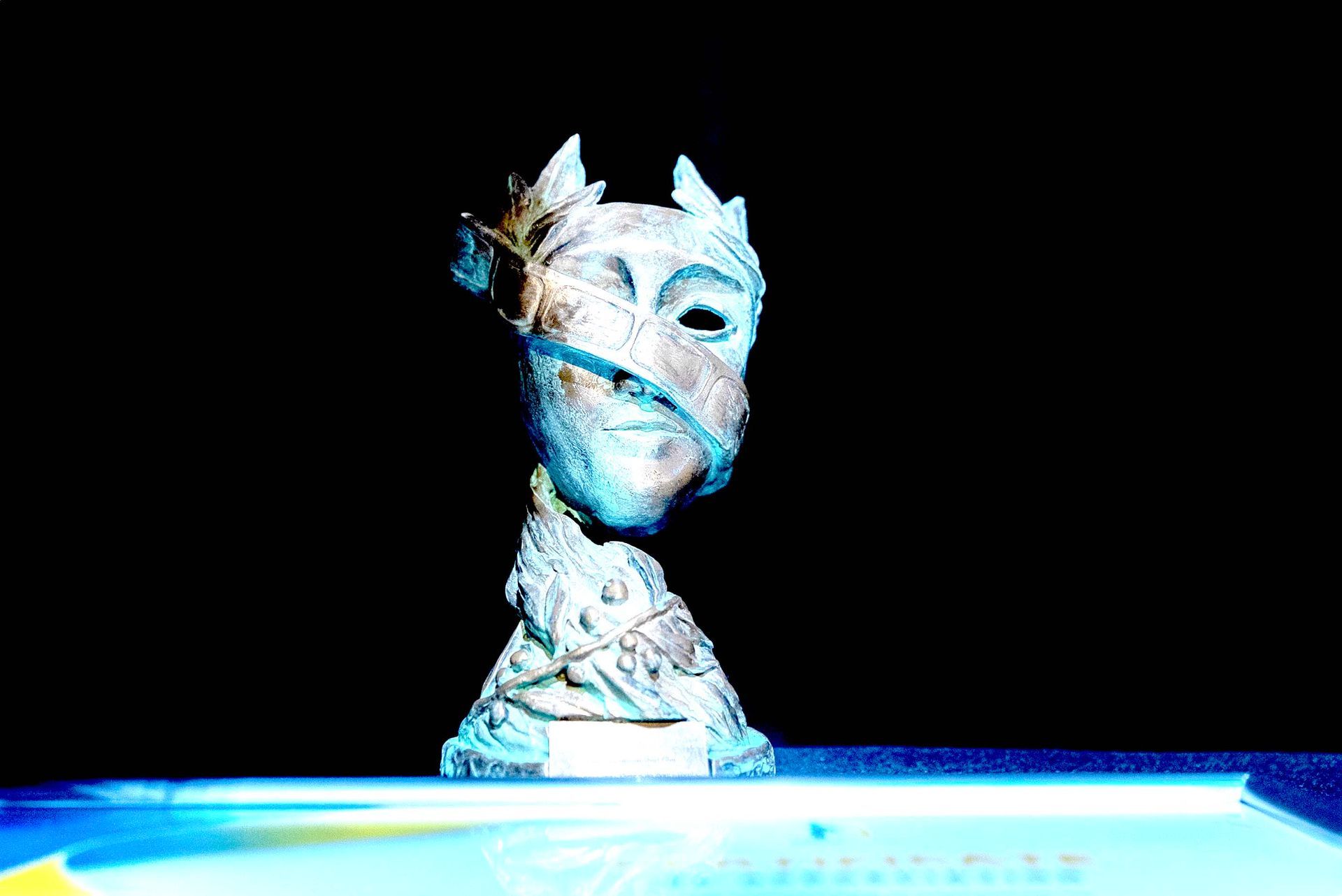 Blue-lit sculpture head with a crown-like top on a black background, displayed above a glowing platform