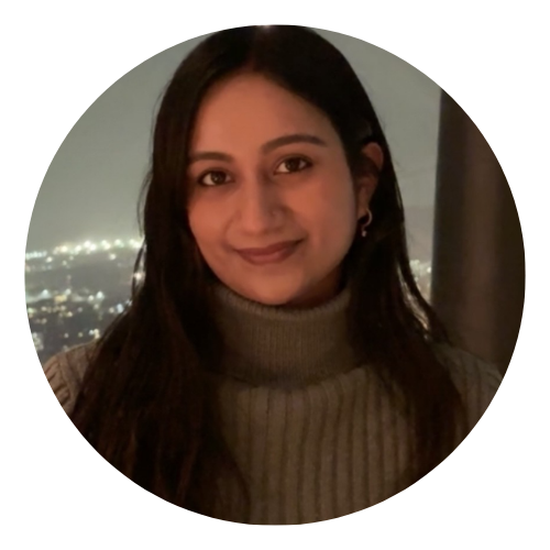 Portrait of a smiling person in a brown turtleneck, with city lights blurred in the background