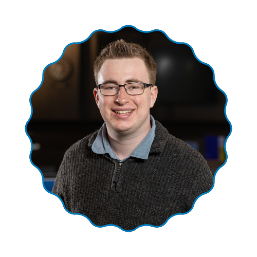 Smiling person wearing glasses and a dark sweater, framed in a scalloped blue-edged circle.