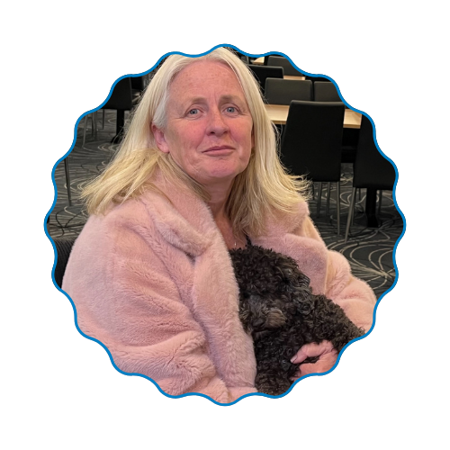Woman in a pink coat holding a small black dog indoors, seated among empty chairs.