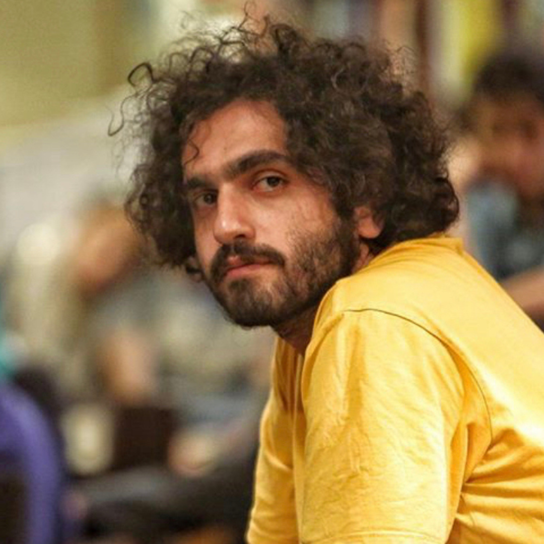 Curly-haired person in a yellow shirt looking toward the camera in a blurred indoor setting
