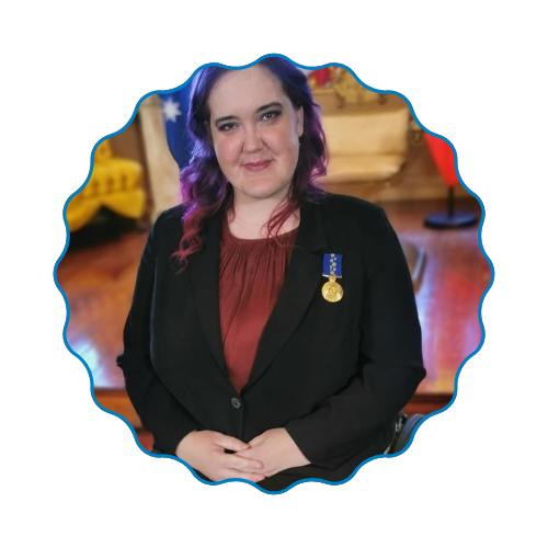 Woman in black blazer with medal, standing indoors against a blurred warm-toned background.