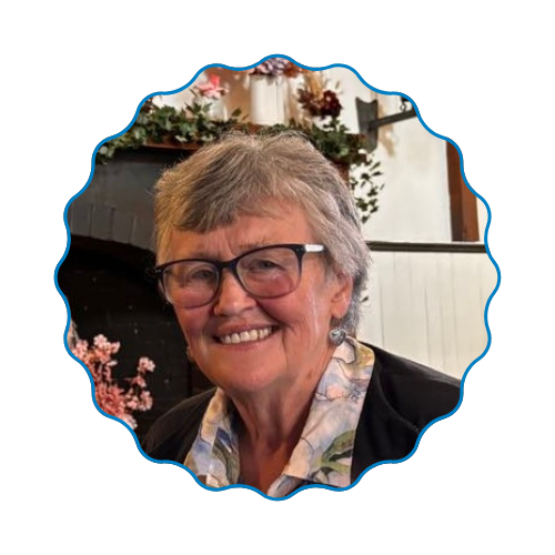 Smiling person with short gray hair and glasses, in a black jacket, seated indoors by floral decor