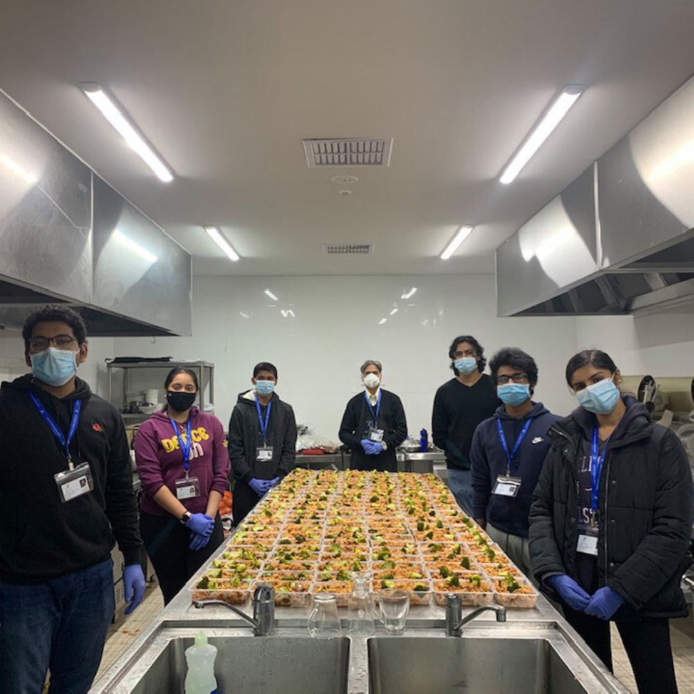 People standing around a long table filled with trays of food in a bright commercial kitchen