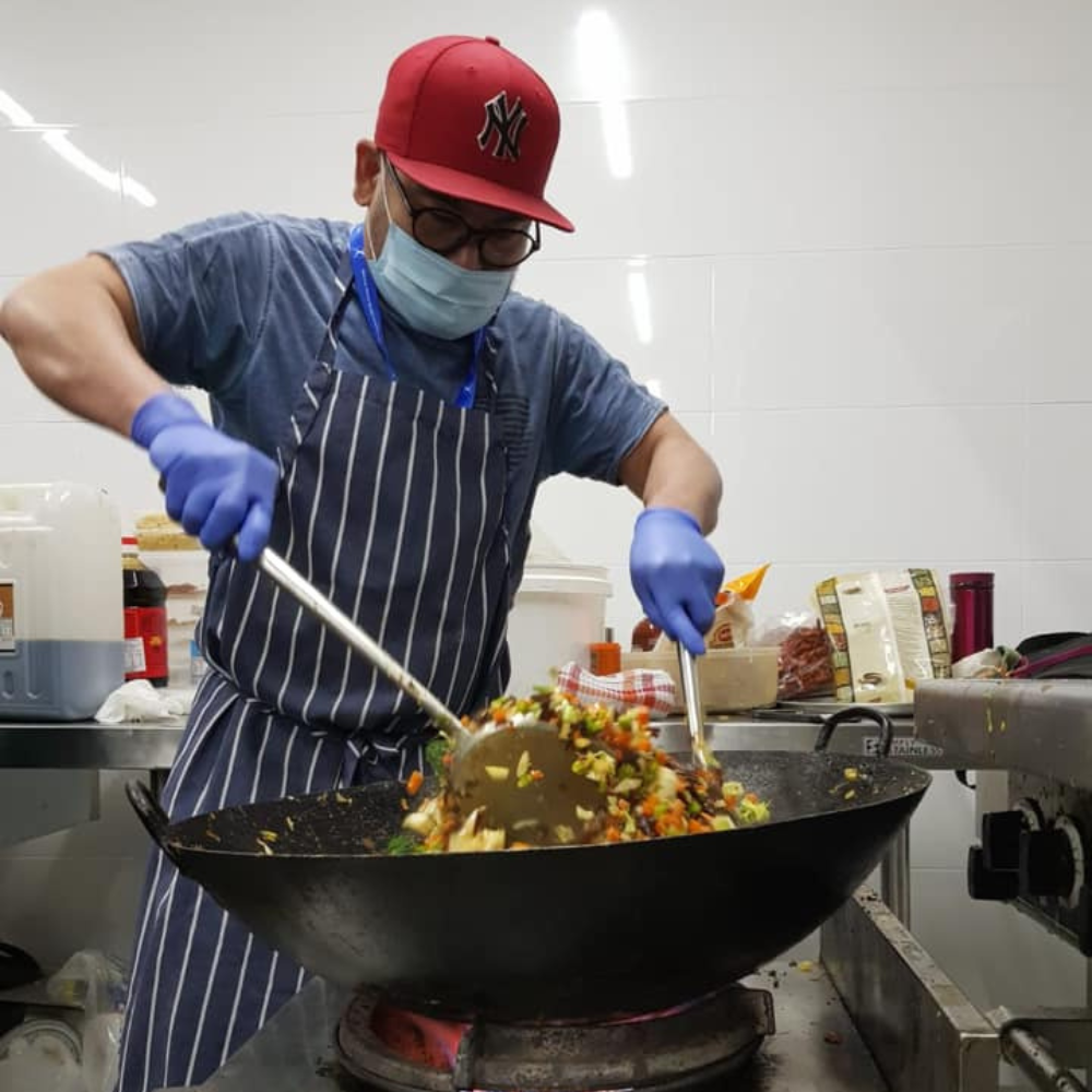 Cook in apron and gloves stirring food in a large wok in a kitchen