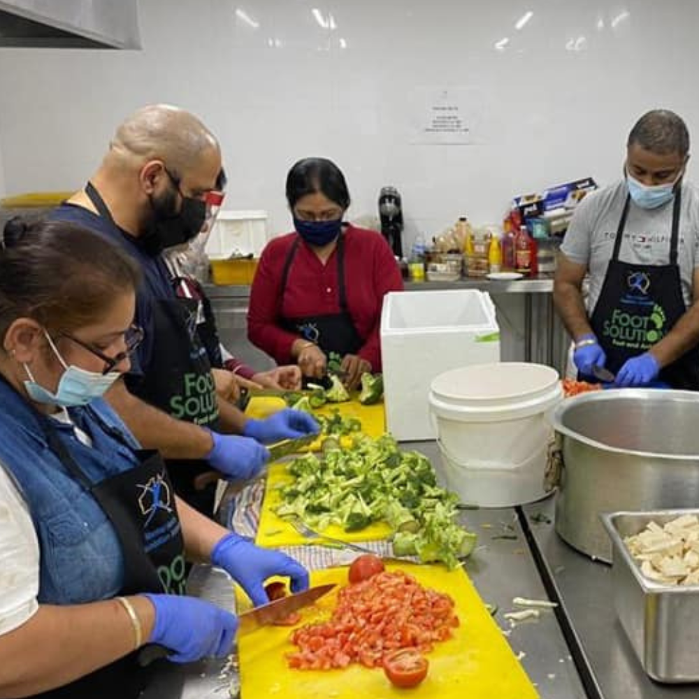 People in gloves and aprons chop vegetables at a commercial kitchen prep table.
