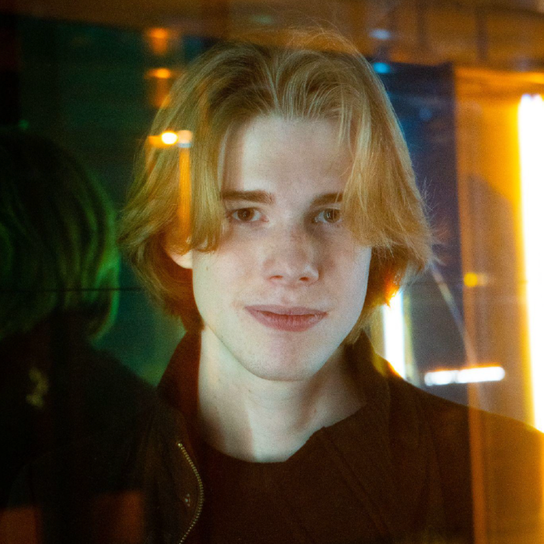 Portrait of a person with blond hair in a dim, neon-lit setting, wearing a black top.