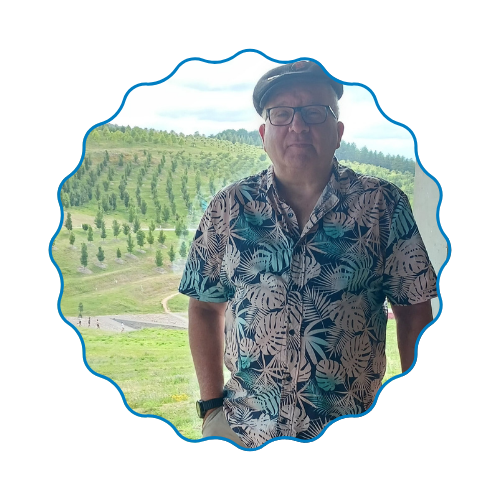 Person in a floral shirt and cap standing outdoors in front of a green hillside and trees.