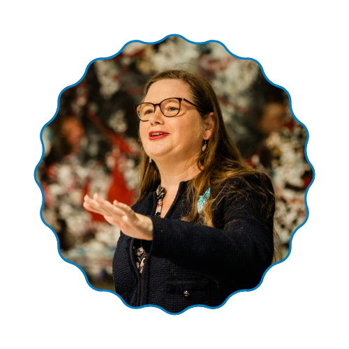 Woman speaking with one hand raised, wearing glasses and a dark jacket, against a blurred floral background.