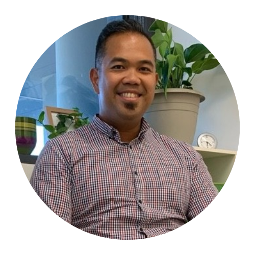 Smiling person in a checkered shirt seated indoors beside a plant and shelves.
