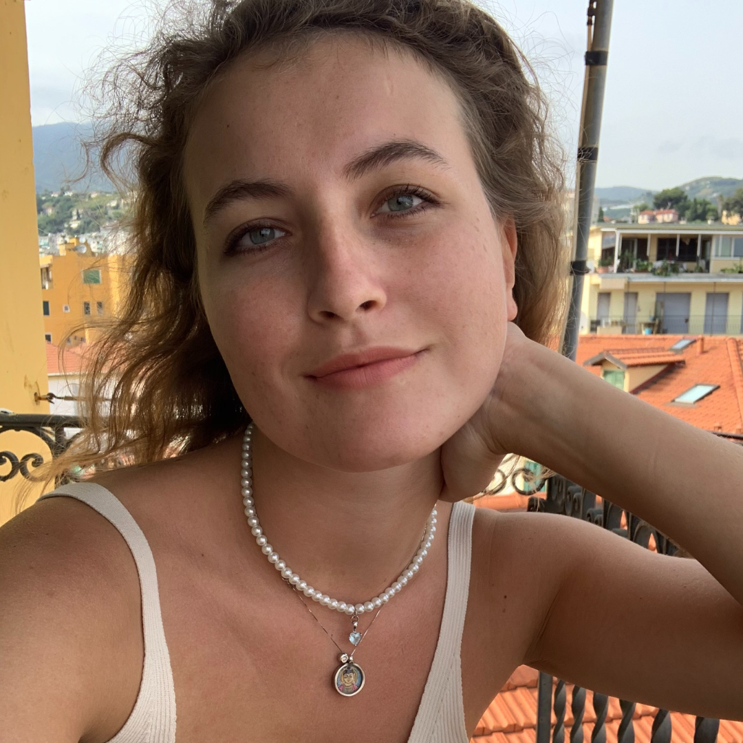 Woman in a white top and pearl necklace posing on a balcony with mountains and rooftops in the background