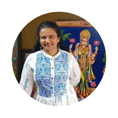 Smiling woman in a white and blue kurta standing indoors beside a framed Hindu deity painting
