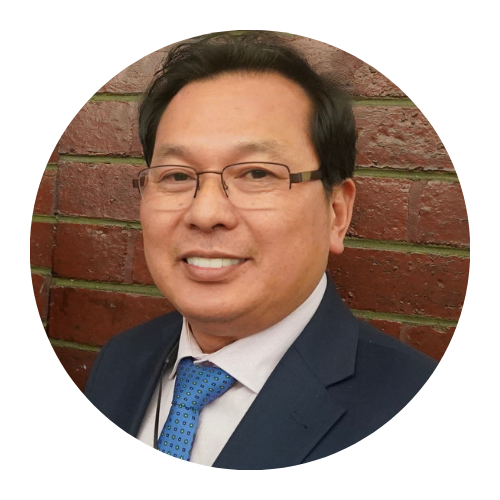 Smiling man in glasses, navy suit, and blue tie against a brick wall.