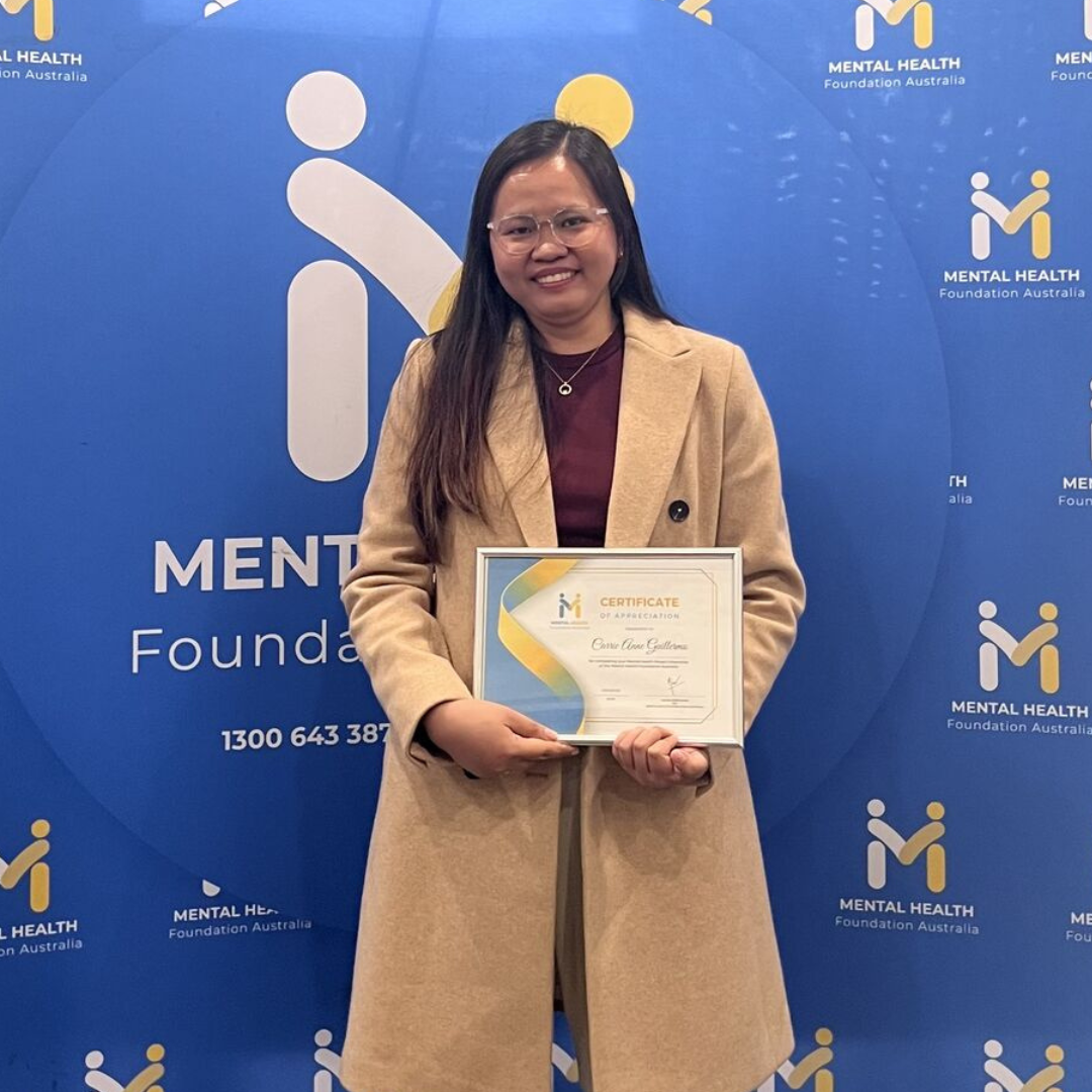 Woman holding a certificate, smiling in front of a blue Mentel Health Foundation backdrop.