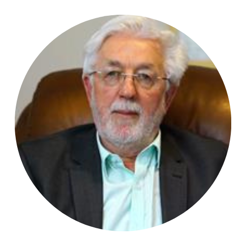 Portrait of a gray-haired man in glasses, wearing a dark blazer and light shirt, seated against a brown chair.