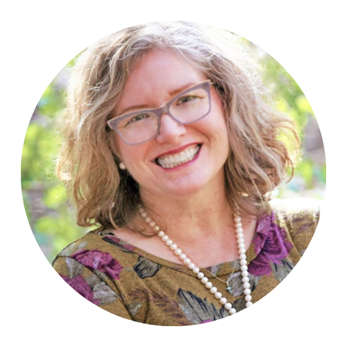 Smiling woman with glasses and curly hair, wearing a floral top and pearl necklace, outdoors in soft light