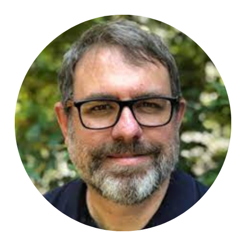 Portrait of a man with glasses and a salt-and-pepper beard, smiling outdoors with blurred greenery behind him