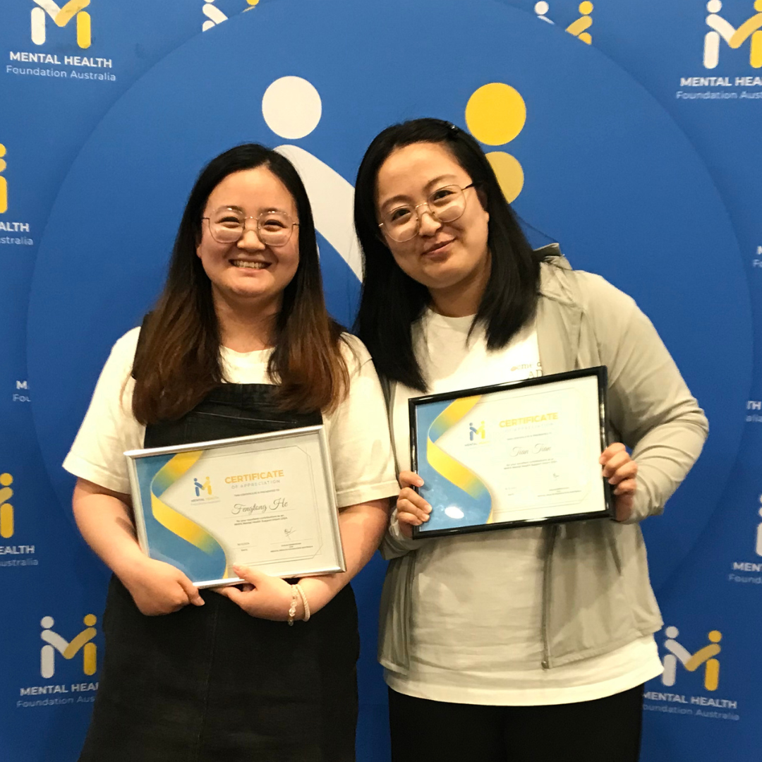 Two people smiling and holding framed certificates in front of a blue event backdrop.