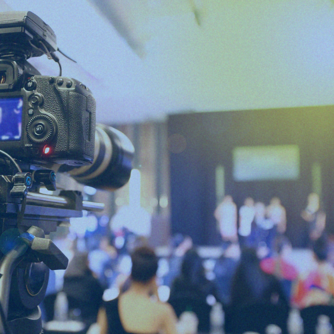 Camera filming a blurred indoor stage presentation with an audience in the foreground