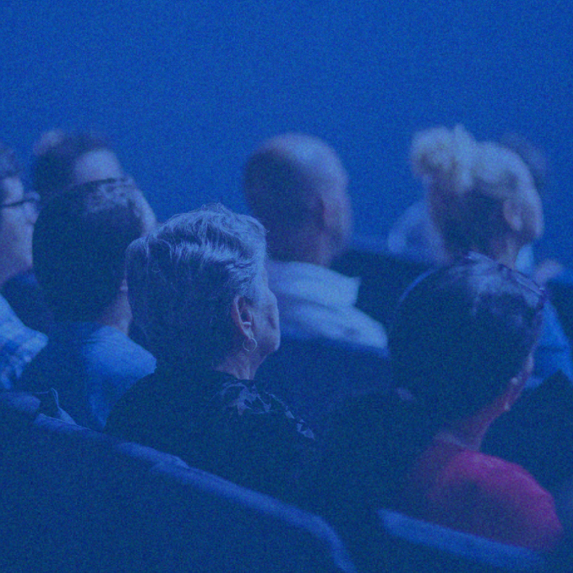 Audience seated in dim blue lighting, viewed from behind during an event