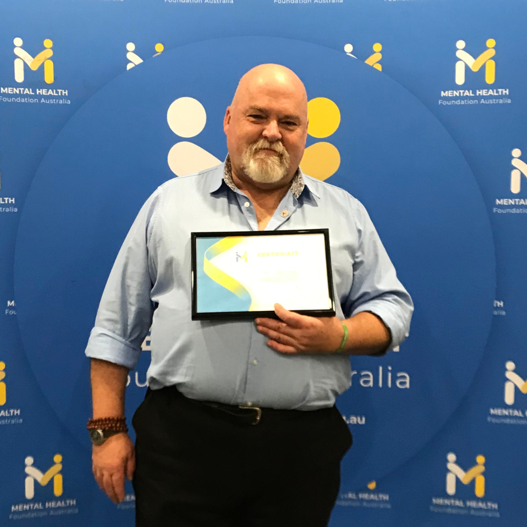 Man holding a framed certificate in front of a blue event backdrop with logos