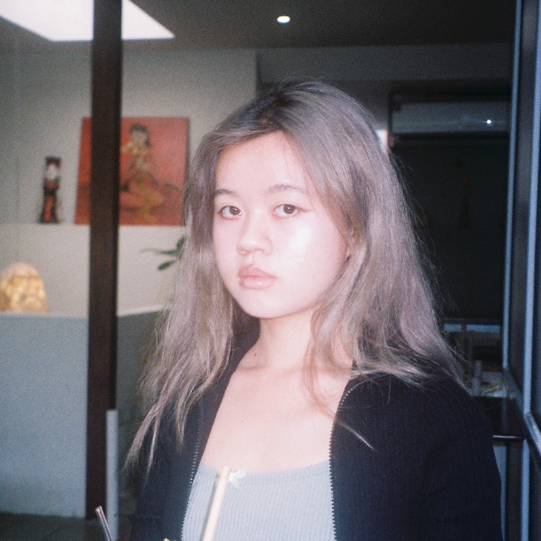Portrait of a person with long hair indoors, wearing a black jacket and gray top, in a softly lit room