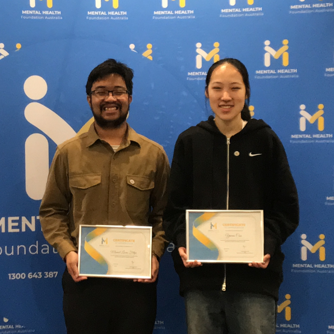 Two people holding certificates in front of a blue mental health backdrop.