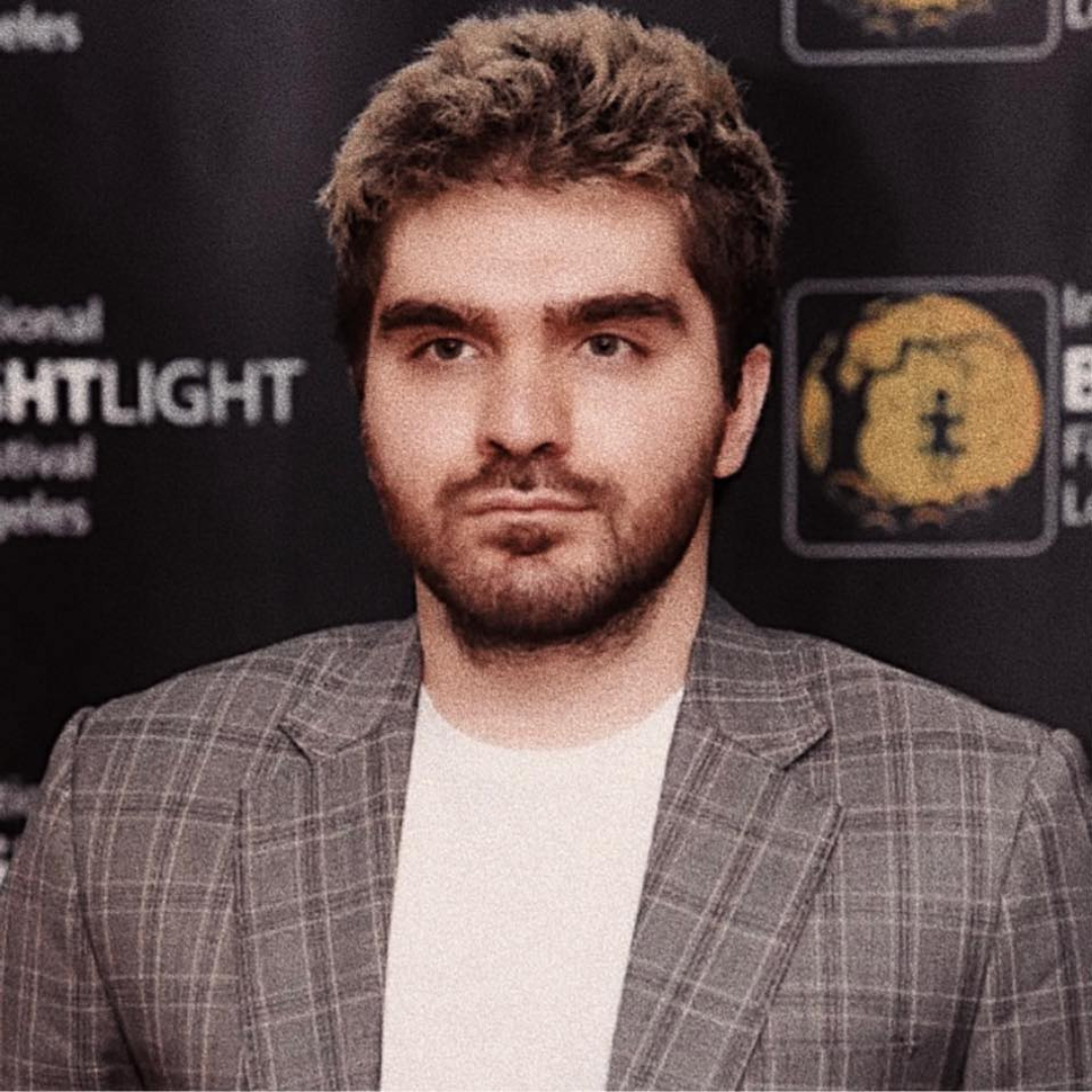 Man in a gray plaid blazer posing at a step-and-repeat event backdrop