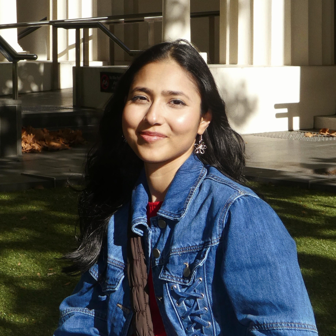 Woman in a blue denim jacket sitting outdoors in sunlight, smiling at the camera.