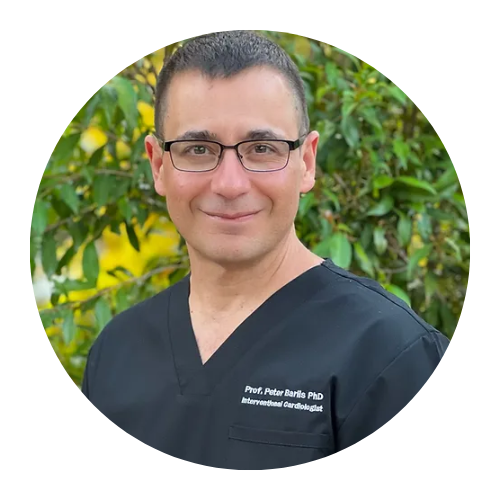 Smiling person in black scrubs, standing outdoors in front of green foliage.