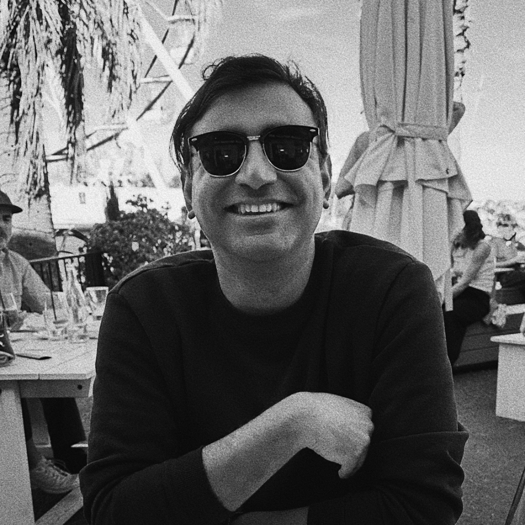 Smiling person in sunglasses seated outdoors at a table, wearing a black shirt.