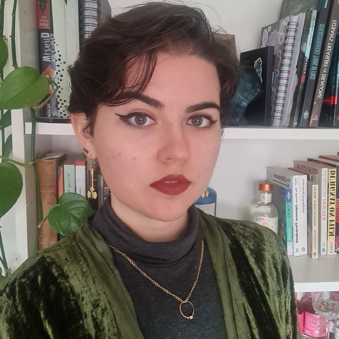 Person with short dark hair and red lipstick, wearing a green velvet top, standing in front of bookshelves.