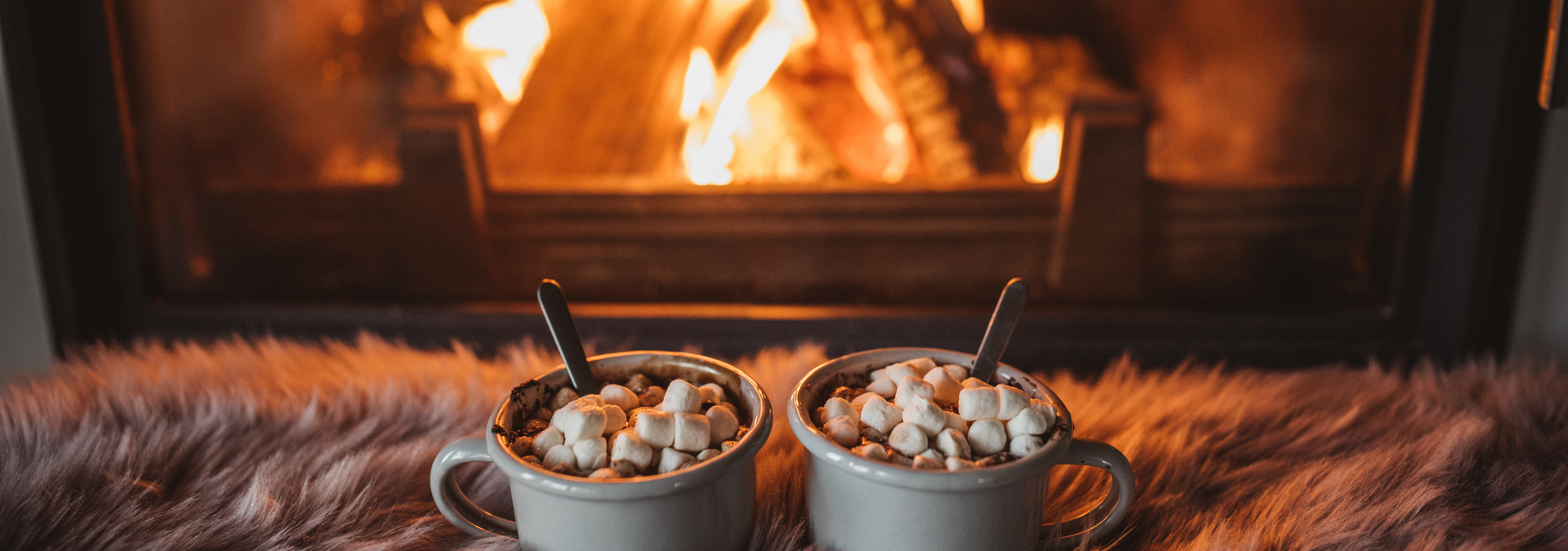 Two mugs of hot cocoa with marshmallows on a fur blanket before a glowing fireplace
