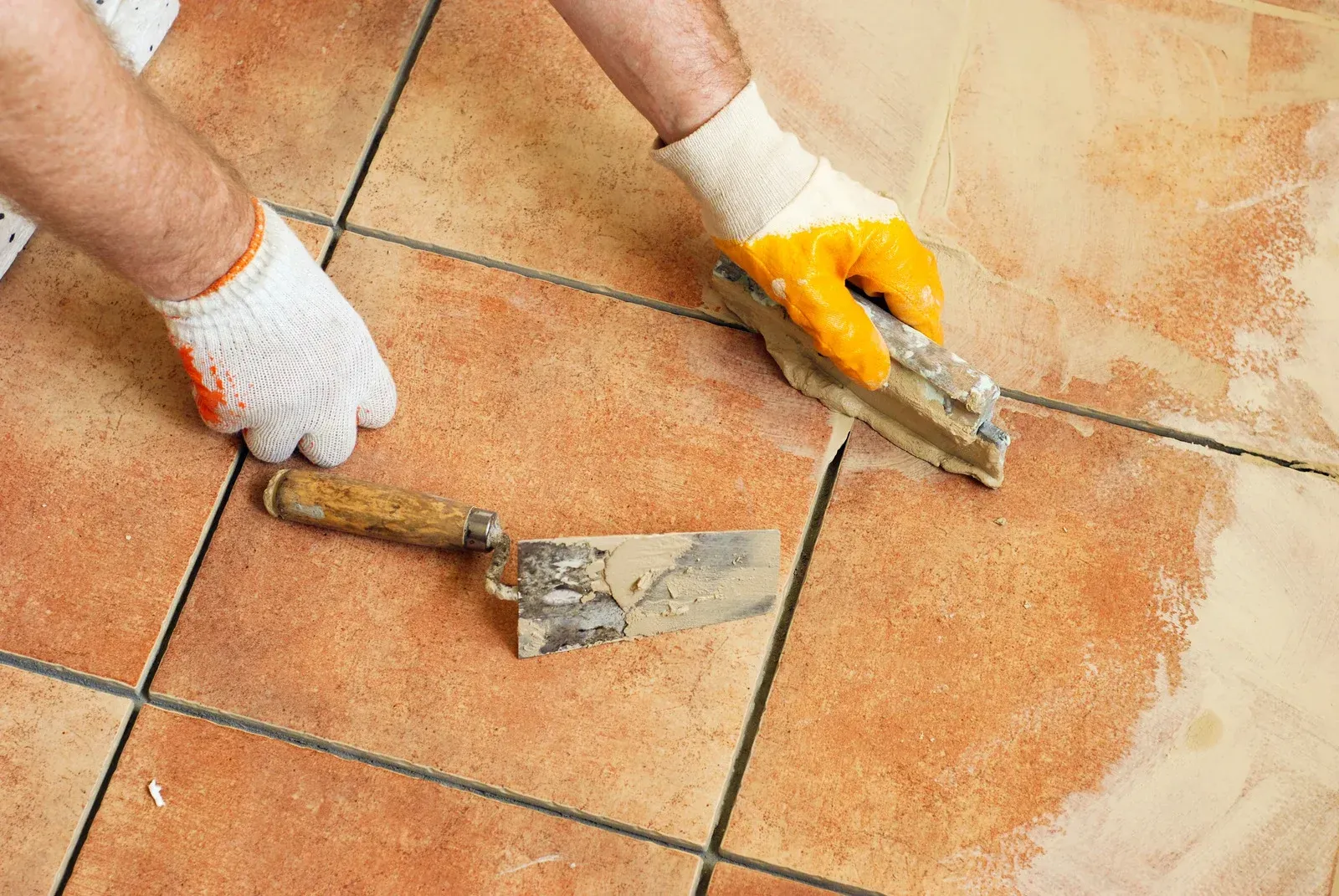 Person tiling floor with a trowel, wearing gloves. Orange tile, tan grout.