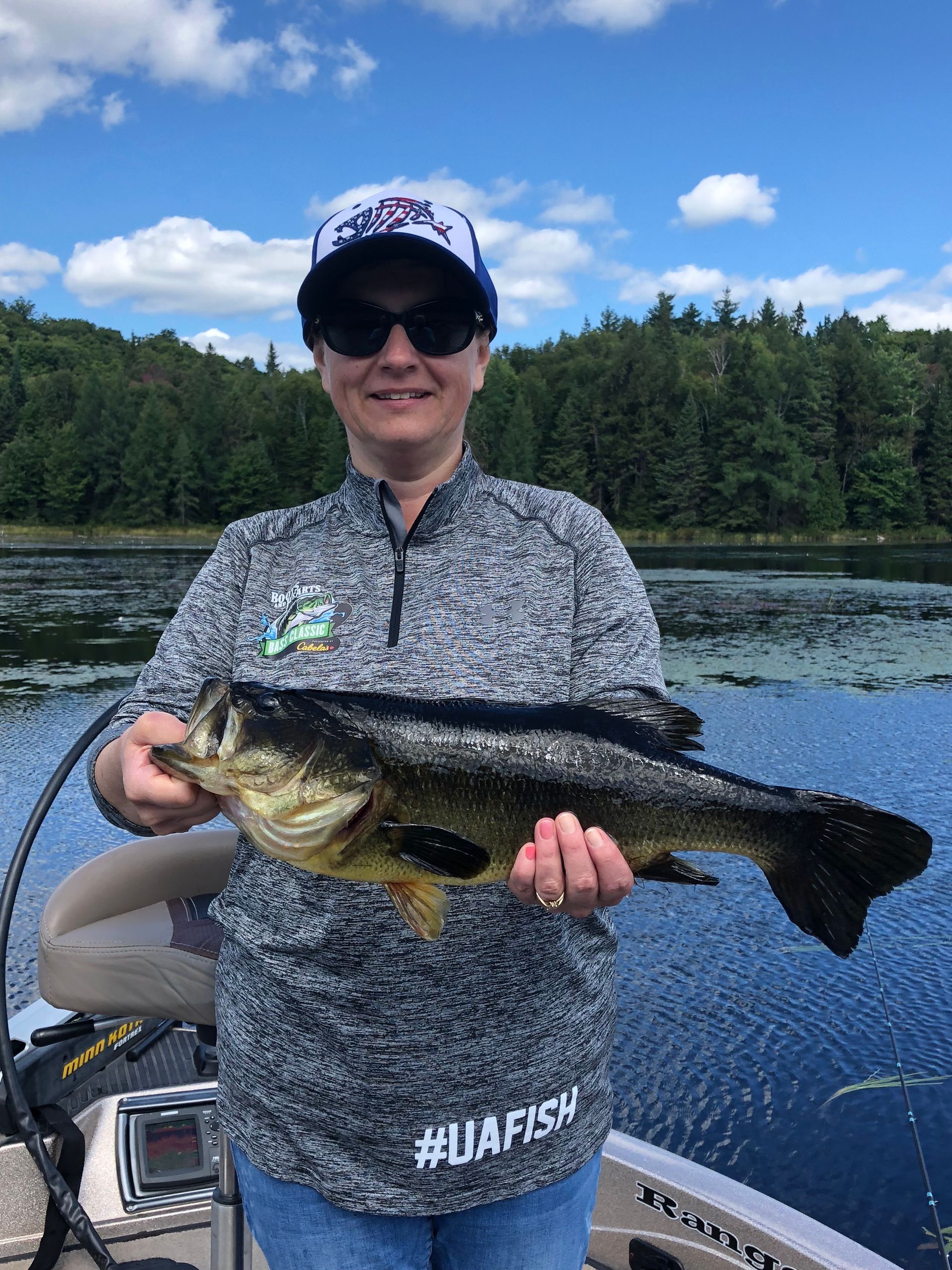 Person holding a large fish beside a lake, with trees and blue sky in the background.