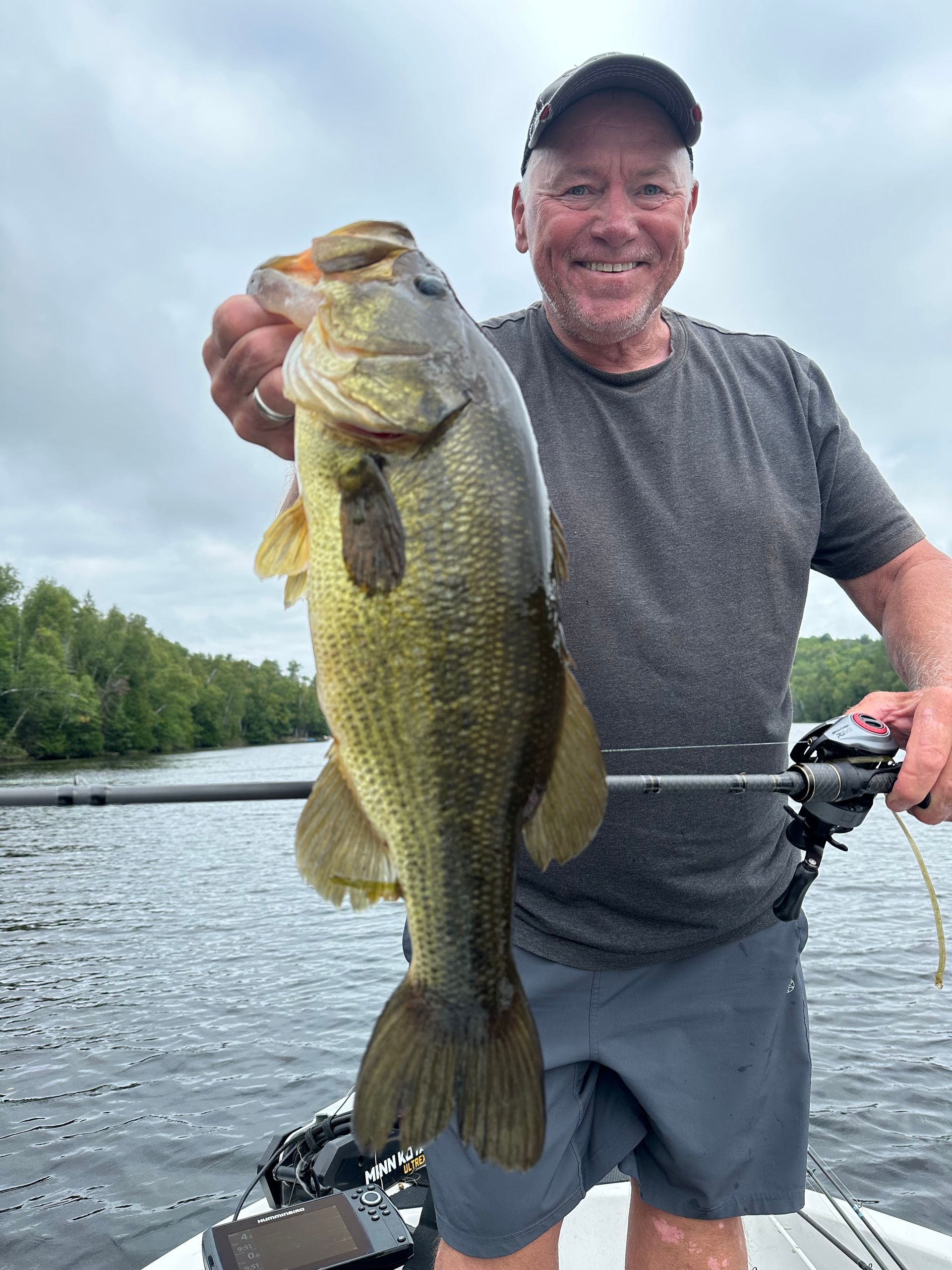 Angler holding a large bass on a lake, smiling beside a fishing rod and reel.