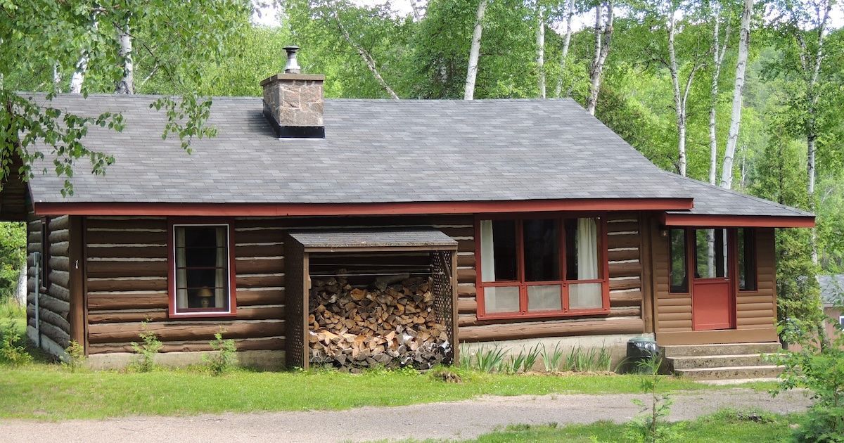 Rustic log cabin with red trim and stacked firewood, surrounded by green trees