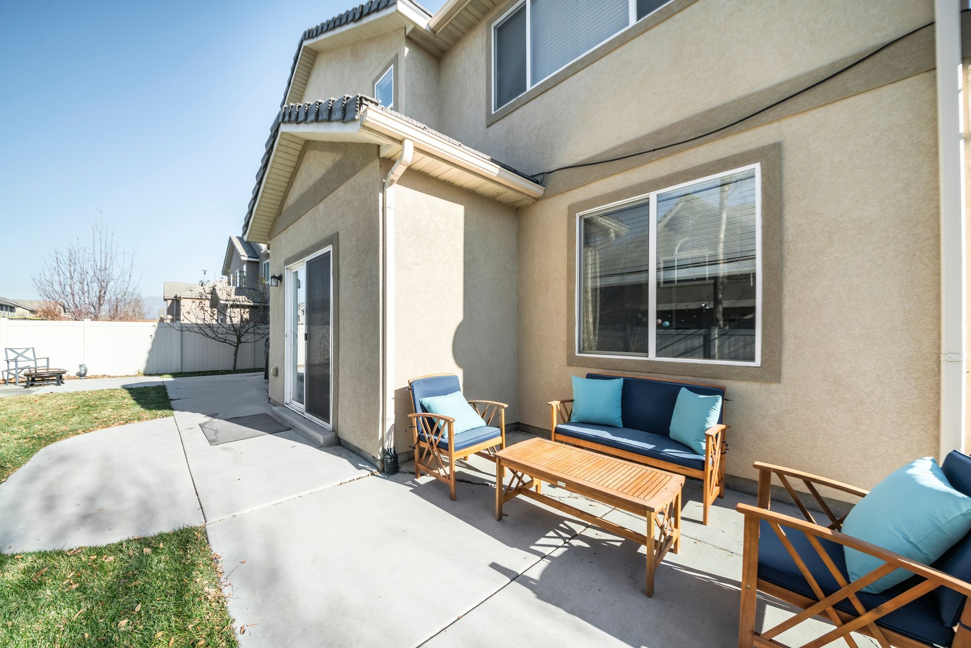 Patio with blue-cushioned chairs and wooden table outside a beige house.