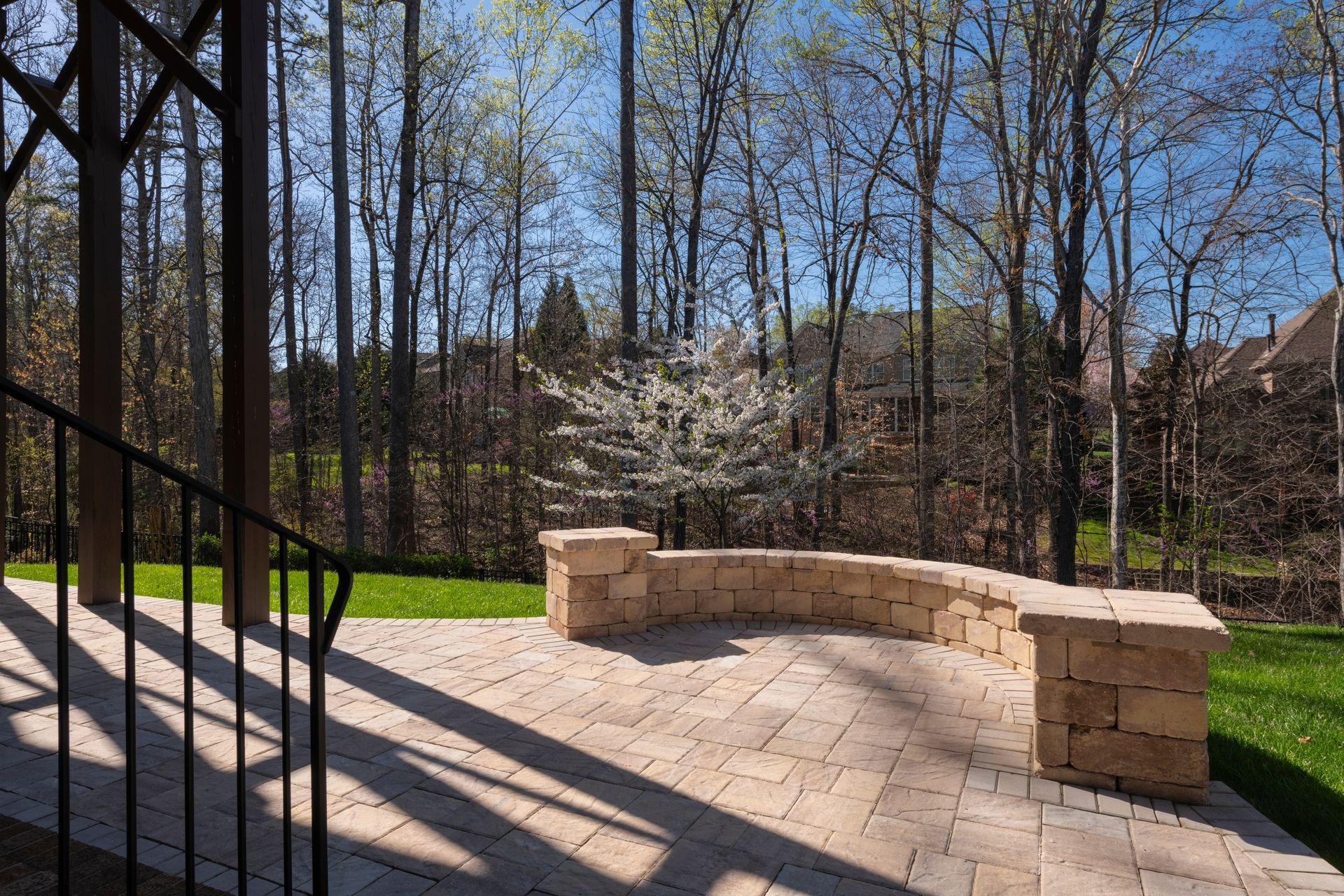 Stone bench on a sunlit patio beside black railings, with trees and a blossoming tree in the background