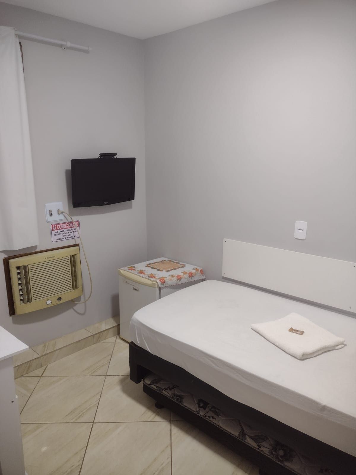 Um quarto de hotel simples com uma cama de solteiro, uma TV de parede, um ar-condicionado de janela e uma pequena geladeira.