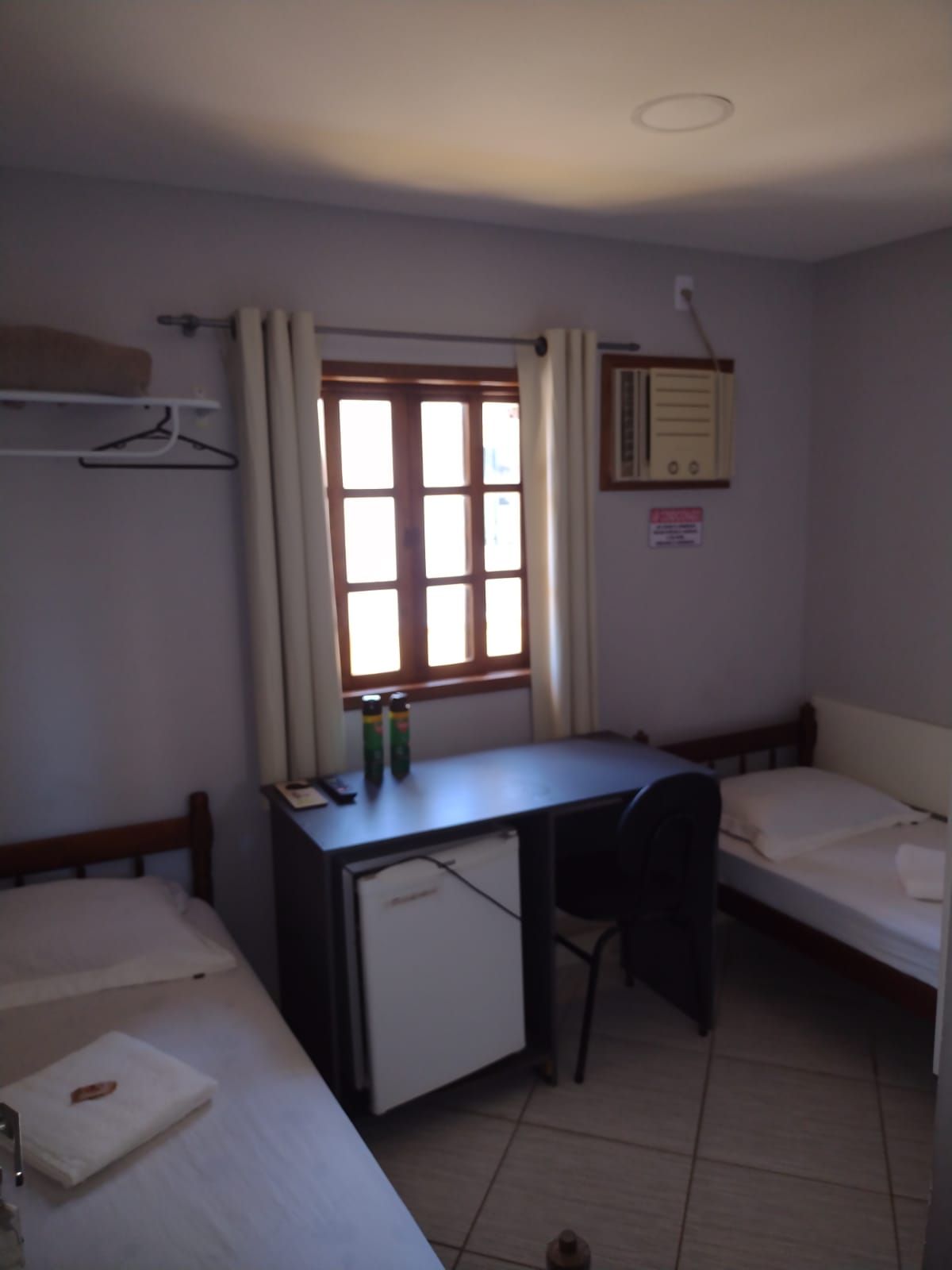 Um pequeno quarto de hotel com duas camas de solteiro, uma escrivaninha com frigobar embaixo, uma cadeira e uma janela com cortinas.