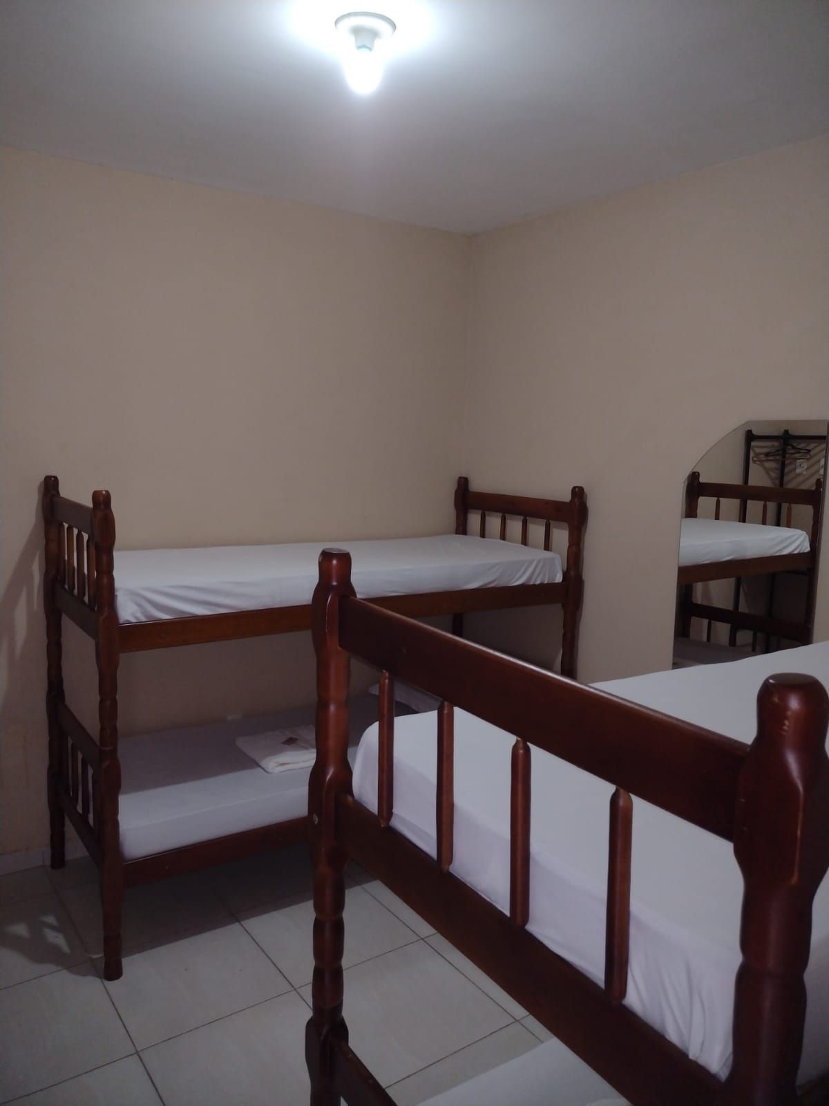 Um pequeno quarto com duas camas de beliche de madeira com lençóis brancos e uma parede bege, iluminado por uma única lâmpada de teto.
