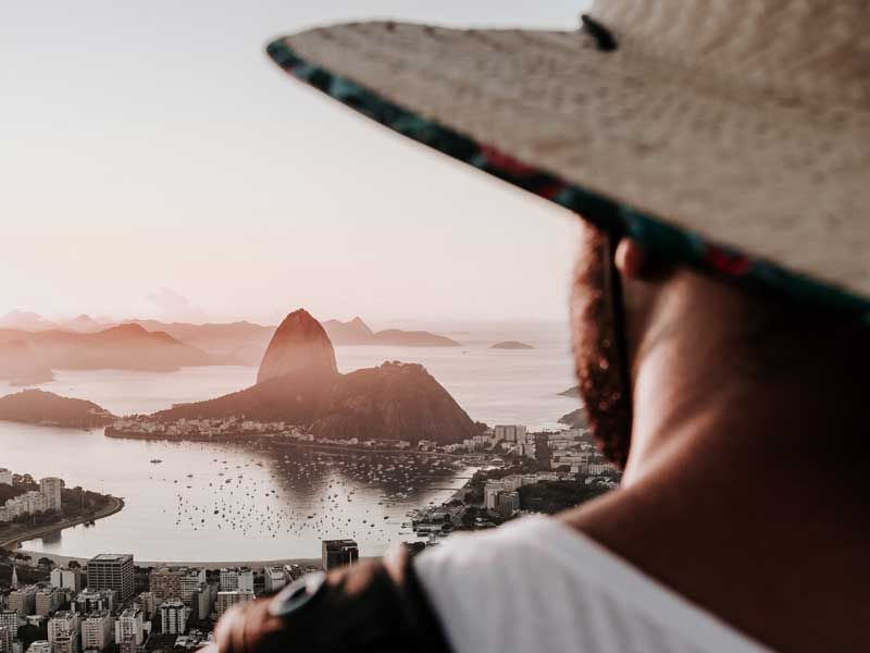 Uma pessoa com um chapéu de palha observa a orla do Rio de Janeiro, com o Pão de Açúcar visível ao longe.