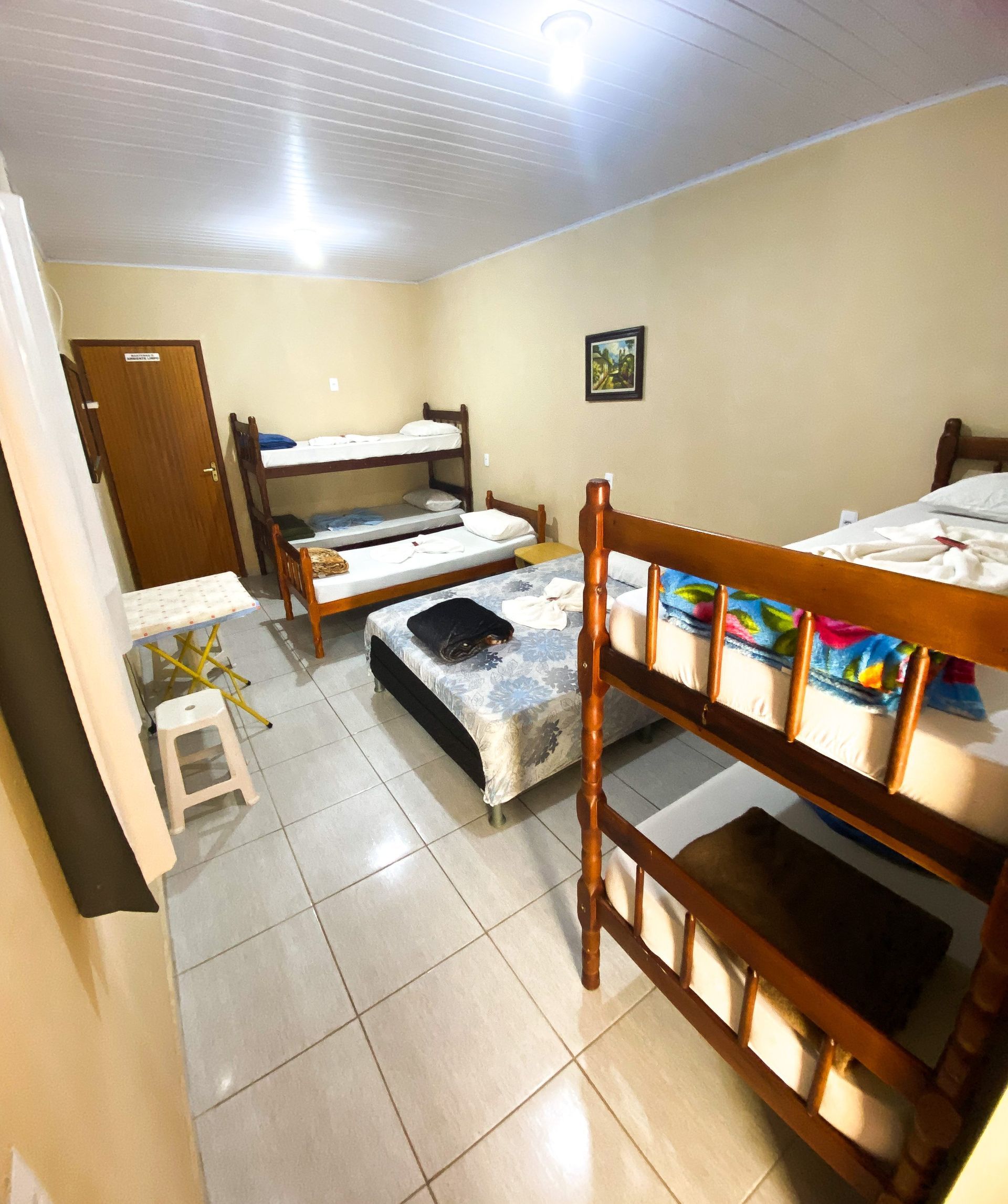 Um quarto de dormitório de albergue com piso de azulejo, duas camas de beliche de madeira, uma cama de solteiro, uma mesinha e um banquinho.