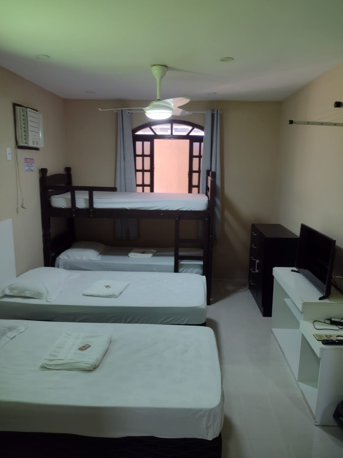 Um quarto de hotel com duas camas de solteiro, um beliche de madeira, uma TV de tela plana em um suporte branco e decoração em tons de bege.