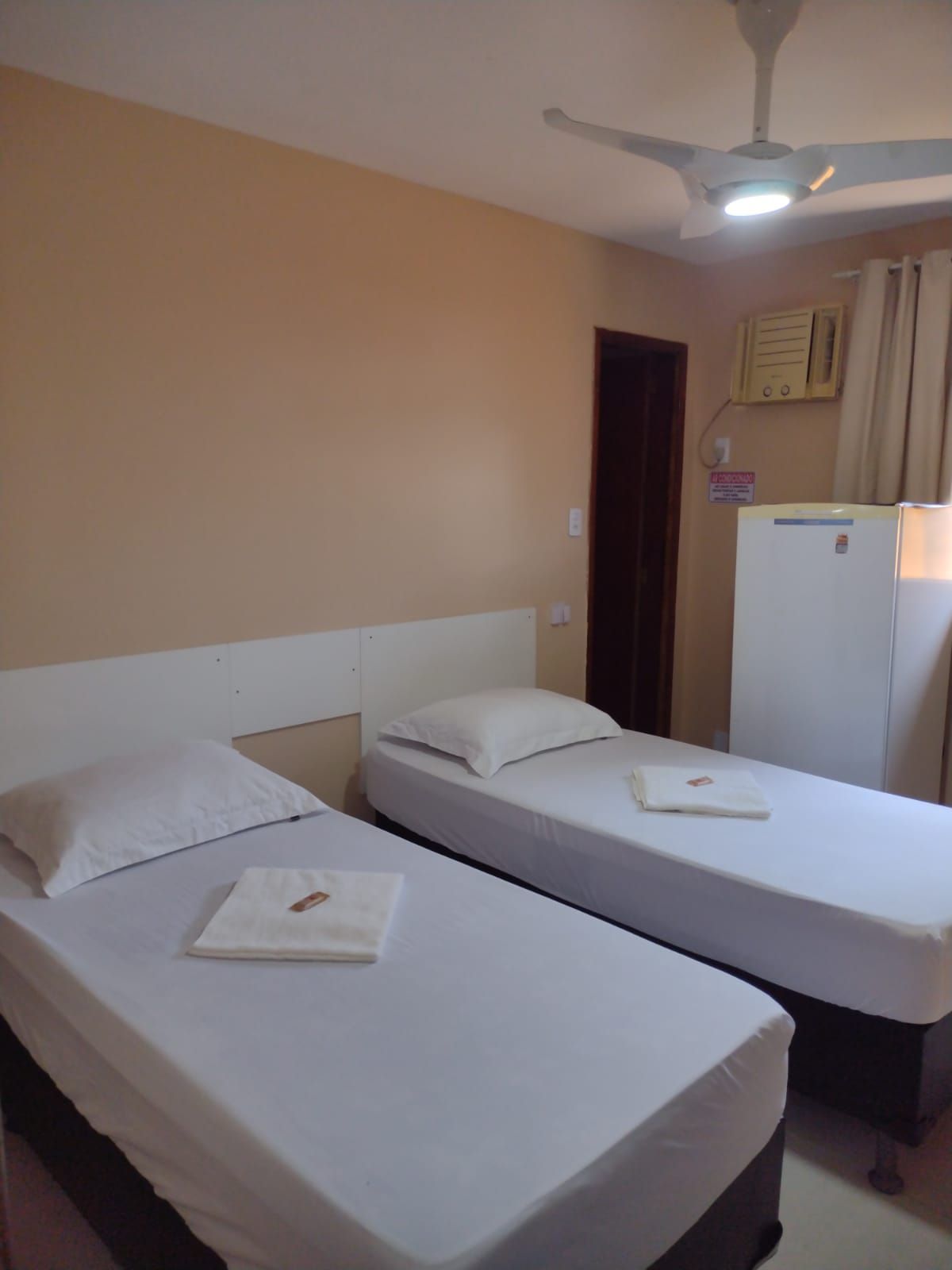 Um quarto de hotel com duas camas de solteiro com lençóis brancos, um ventilador de teto e um frigobar perto de uma janela com cortina.