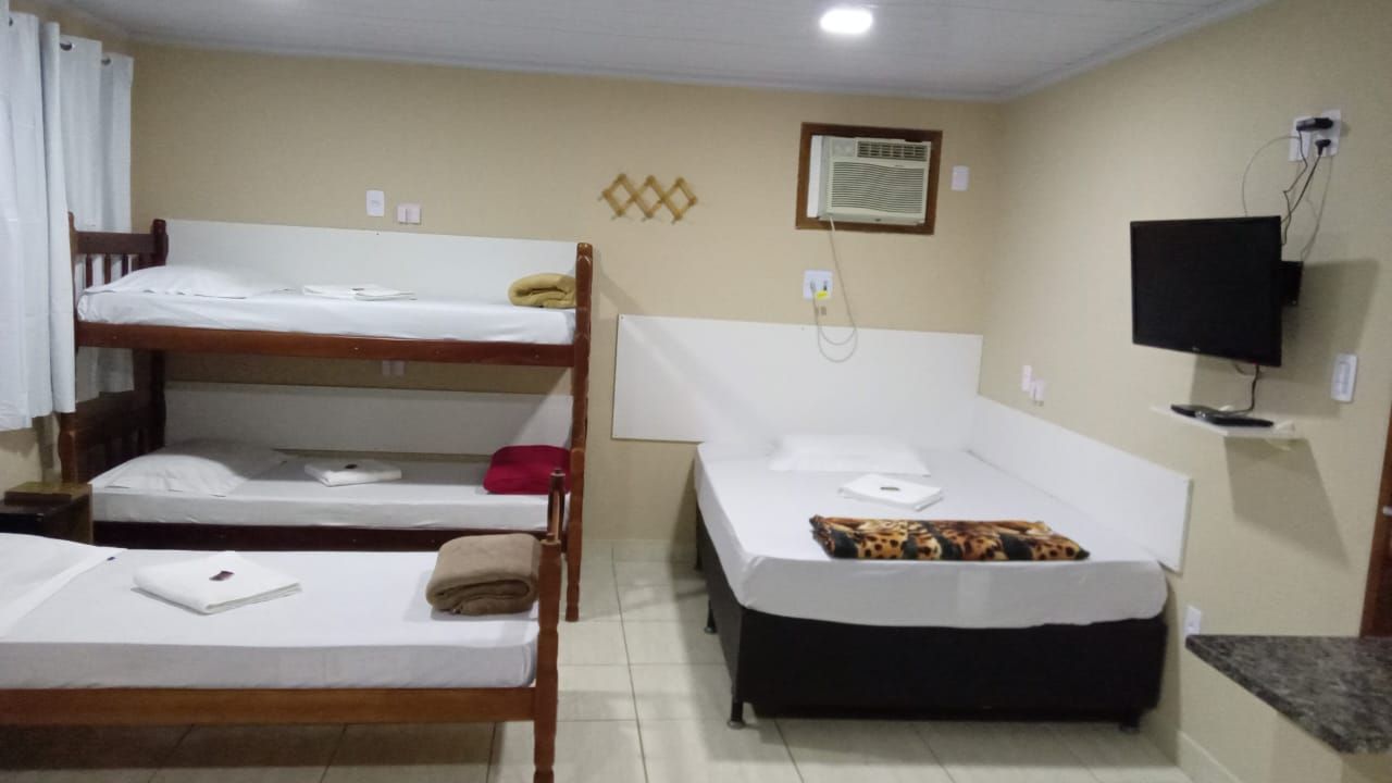Um quarto de hotel com um beliche de madeira, uma cama de solteiro e uma cama de casal, com TV de parede e paredes em tons neutros.
